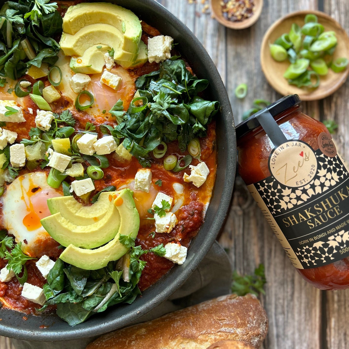  bowl of Shakshuka with avocado, feta, and greens on a wooden table with a jar of Shakshuka Sauce.