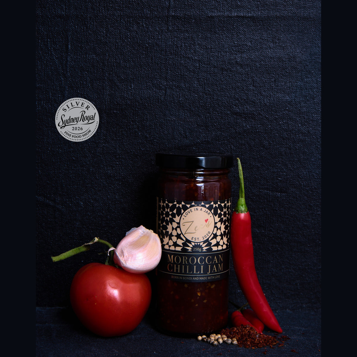 A glass jar of Moroccan Chilli Jam from Sydney Royal is styled with a red tomato, a garlic bulb, a whole red chili pepper, and spices on a dark textured background.