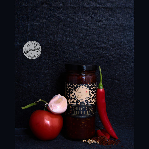 A glass jar of Moroccan Chilli Jam from Sydney Royal is styled with a red tomato, a garlic bulb, a whole red chili pepper, and spices on a dark textured background.