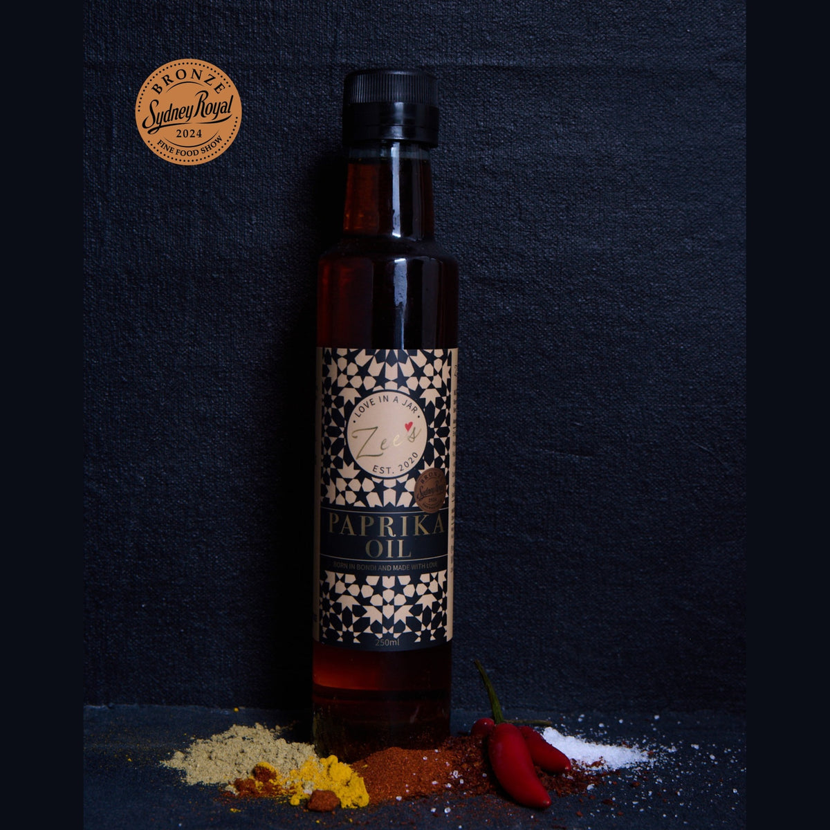 A bottle of Zee's Paprika Oil stands on a dark textured surface with spices and red chili peppers scattered around it. A circular 'Bronze Sydney Royal award 2024' award seal is visible in the upper left corner.