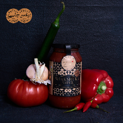 A jar of Fronz Shakshuka Sauce styled with fresh ingredients including a red tomato, garlic, red bell pepper, and chili peppers on a dark textured background.
