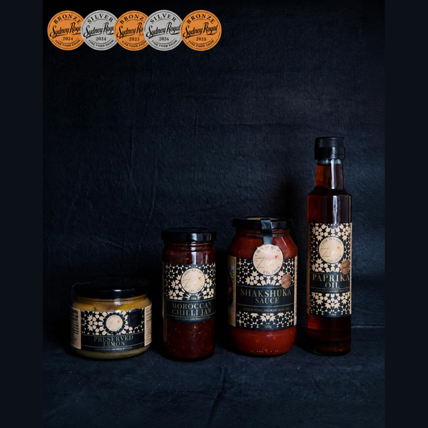 Four jars and one tall bottle of Paprika oil from Zee's love in a Jar, arranged against a dark textured background. The products include Shakshuka sauce, Moroccan chilli jam, Preserved lemon, and various flavored sauces with black and gold labels.